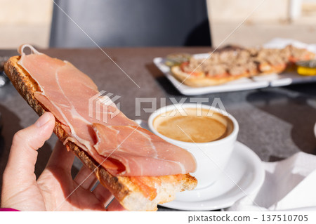 Woman eating tostada with ham and drinking coffee with milk in a cafe outside. Spanish breakfast. 137510705