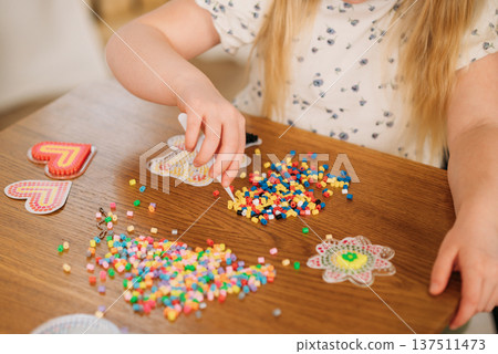 Child's hands creating a bear shape using colorful fuse beads on a plastic pegboard at a wooden table. thermomosaic Child's hands creating a bear shape using colorful fuse beads on a plastic pegboard at a wooden table. thermomosaic 137511473