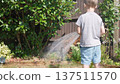 Funny little boy watering lawn plants in garden housing backyard. Adorable child playing with irrigation hose at hot sunny summer outdoors. Children help with housework. activity for kids. Childhood 137511570