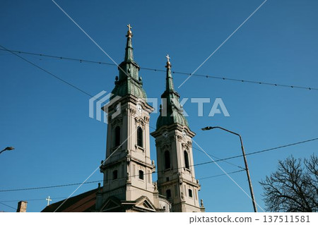 Twin towers of a historic Orthodox church rising above the city skyline in Pancevo, Serbia. Architectural landmark with crosses, clocks and baroque details under clear blue sky Twin towers of a historic Orthodox church rising above the city skyline in Pancevo, Serbia. Architectural landmark with crosses, clocks and baroque details under clear blue sky 137511581