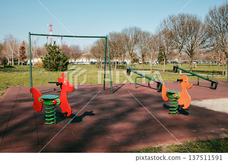 Colorful playground equipment with spring riders, swing and seesaw in a sunny park. Outdoor children recreation area with bright play structures and rubber safety surface Colorful playground equipment with spring riders, swing and seesaw in a sunny park. Outdoor children recreation area with bright play structures and rubber safety surface 137511591