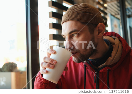 Young man in red jacket drinking carbonated soda from paper cup while sitting near window in fast food restaurant. Concept of quick meal, urban lifestyle and casual dining Young man in red jacket drinking carbonated soda from paper cup while sitting near window in fast food restaurant. Concept of quick meal, urban lifestyle and casual dining 137511616