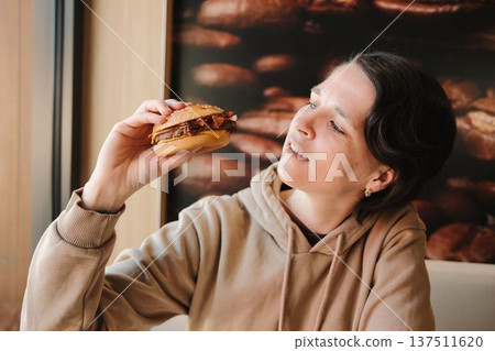Young woman holding a beef burger with bacon on a sesame bun while sitting in a fast food restaurant and looking out the window. Concept of casual dining and urban lifestyle 137511620