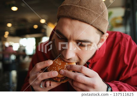Close up of a young man biting a juicy beef burger with bacon on a sesame bun while sitting in a fast food restaurant. Concept of casual dining, street food and quick urban meal Close up of a young man biting a juicy beef burger with bacon on a sesame bun while sitting in a fast food restaurant. Concept of casual dining, street food and quick urban meal 137511625
