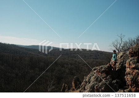 Female traveler standing on rocky cliff Orlove Stene in Fruska Gora, Serbia with German and Australian Shepherd dogs overlooking vast forest valley. Adventure and freedom concept in nature 137511658