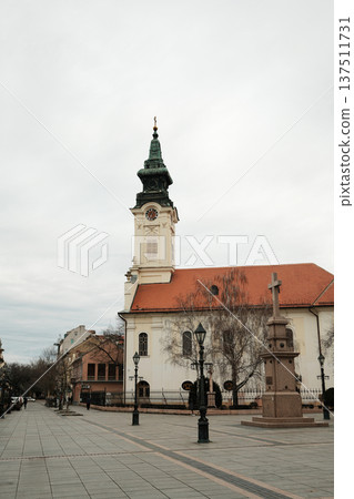 Church with clock tower and stone cross monument on open square in Sombor, Serbia. European religious heritage and city landmark concept 137511731