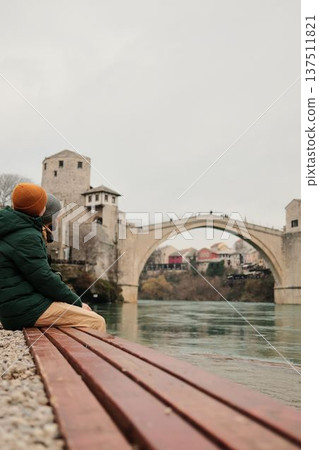 Couple sitting on a wooden bench by the river facing the historic bridge in Mostar. Slow travel and peaceful reflection concept connected with cultural heritage and urban nature Couple sitting on a wooden bench by the river facing the historic bridge in Mostar. Slow travel and peaceful reflection concept connected with cultural heritage and urban nature 137511821