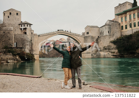 Smiling couple with open arms standing by the river near the historic bridge in Mostar. Travel freedom and shared happiness concept inspired by exploration and cultural discovery Smiling couple with open arms standing by the river near the historic bridge in Mostar. Travel freedom and shared happiness concept inspired by exploration and cultural discovery 137511822