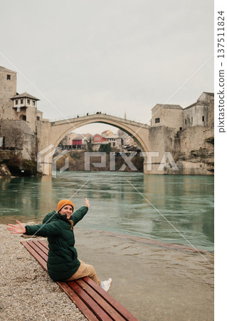 A woman sits on a wooden bench by the river with arms open facing the Old Bridge in Mostar. Concept of freedom travel joy and personal connection with historic places A woman sits on a wooden bench by the river with arms open facing the Old Bridge in Mostar. Concept of freedom travel joy and personal connection with historic places 137511824