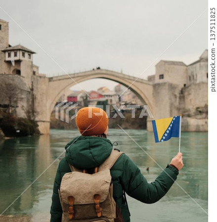 A traveler with a backpack holds the Bosnia and Herzegovina flag while facing the historic bridge across the river. Concept of adventure travel, freedom, cultural discovery, and personal exploration A traveler with a backpack holds the Bosnia and Herzegovina flag while facing the historic bridge across the river. Concept of adventure travel, freedom, cultural discovery, and personal exploration 137511825