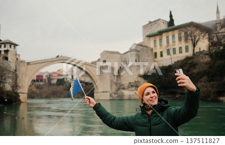 A woman takes a selfie holding the Bosnia and Herzegovina flag near the Old Bridge by the river. Concept of modern travel storytelling, social media culture, tourism lifestyle, and personal memories 137511827