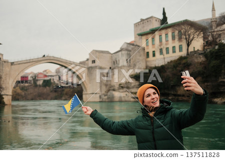 A woman takes a selfie holding the Bosnia and Herzegovina flag near the Old Bridge by the river. Concept of modern travel storytelling, social media culture, tourism lifestyle, and personal memories A woman takes a selfie holding the Bosnia and Herzegovina flag near the Old Bridge by the river. Concept of modern travel storytelling, social media culture, tourism lifestyle, and personal memories 137511828