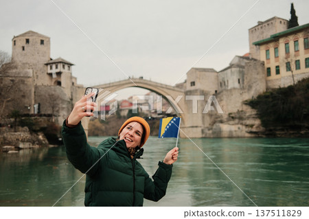 A woman takes a selfie holding the Bosnia and Herzegovina flag near the Old Bridge by the river. Concept of modern travel storytelling, social media culture, tourism lifestyle, and personal memories A woman takes a selfie holding the Bosnia and Herzegovina flag near the Old Bridge by the river. Concept of modern travel storytelling, social media culture, tourism lifestyle, and personal memories 137511829
