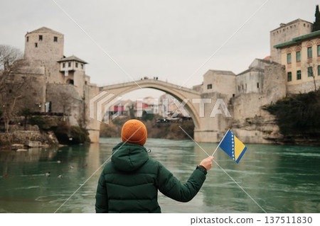 A woman seen from behind holds the Bosnia and Herzegovina flag while looking at the Old Bridge over the river. Concept of reflection, travel inspiration, personal journey, and cultural connection A woman seen from behind holds the Bosnia and Herzegovina flag while looking at the Old Bridge over the river. Concept of reflection, travel inspiration, personal journey, and cultural connection 137511830