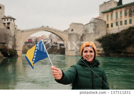 A smiling woman holds the Bosnia and Herzegovina flag in front of the historic stone bridge by the river. Concept of travel memories, national identity, tourism lifestyle, and cultural exploration A smiling woman holds the Bosnia and Herzegovina flag in front of the historic stone bridge by the river. Concept of travel memories, national identity, tourism lifestyle, and cultural exploration 137511831