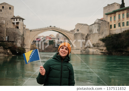A smiling woman holds the Bosnia and Herzegovina flag in front of the historic stone bridge by the river. Concept of travel memories, national identity, tourism lifestyle, and cultural exploration A smiling woman holds the Bosnia and Herzegovina flag in front of the historic stone bridge by the river. Concept of travel memories, national identity, tourism lifestyle, and cultural exploration 137511832