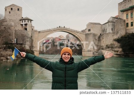 A woman stands by the river holding the Bosnia and Herzegovina flag with arms wide open near the Old Bridge. Concept of freedom, travel identity, cultural pride, and emotional connection with heritage A woman stands by the river holding the Bosnia and Herzegovina flag with arms wide open near the Old Bridge. Concept of freedom, travel identity, cultural pride, and emotional connection with heritage 137511833