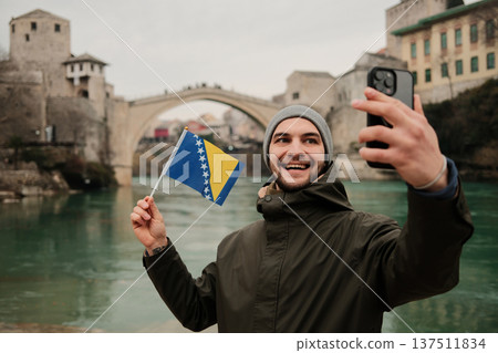 Happy traveler taking a selfie while holding the Bosnia flag near a famous old bridge in Mostar and Neretva river. Concept of tourism personal travel memory and Balkan culture 137511834