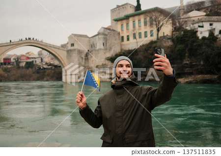 Happy traveler taking a selfie while holding the Bosnia flag near a famous old bridge in Mostar and Neretva river. Concept of tourism personal travel memory and Balkan culture Happy traveler taking a selfie while holding the Bosnia flag near a famous old bridge in Mostar and Neretva river. Concept of tourism personal travel memory and Balkan culture 137511835