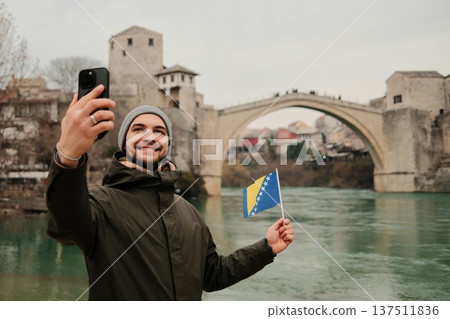Happy traveler taking a selfie while holding the Bosnia flag near a famous old bridge in Mostar and Neretva river. Concept of tourism personal travel memory and Balkan culture 137511836