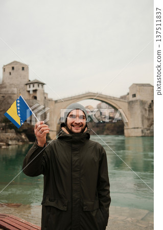 Smiling man holding the Bosnia flag on a riverbank with the historic stone bridge in the background. Travel concept about identity tourism and Balkan heritage Smiling man holding the Bosnia flag on a riverbank with the historic stone bridge in the background. Travel concept about identity tourism and Balkan heritage 137511837