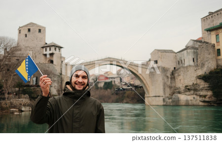 Smiling man holding the Bosnia flag on a riverbank with the historic stone bridge in the background. Travel concept about identity tourism and Balkan heritage Smiling man holding the Bosnia flag on a riverbank with the historic stone bridge in the background. Travel concept about identity tourism and Balkan heritage 137511838