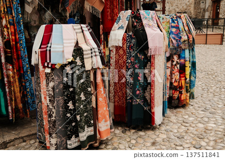 Colorful scarves and shawls hanging at an outdoor market stall on a cobblestone street. Fashion accessories with floral and abstract patterns in a historic setting Colorful scarves and shawls hanging at an outdoor market stall on a cobblestone street. Fashion accessories with floral and abstract patterns in a historic setting 137511841