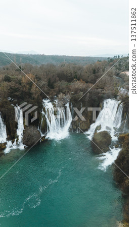 Aerial vertical view of Kravica Waterfalls in Bosnia during winter, with powerful cascades flowing into a turquoise pool, surrounded by bare trees and rocky cliffs in a serene natural landscape Aerial vertical view of Kravica Waterfalls in Bosnia during winter, with powerful cascades flowing into a turquoise pool, surrounded by bare trees and rocky cliffs in a serene natural landscape 137511862