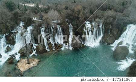Aerial horizontal view of Kravica Waterfalls in Bosnia during winter, with powerful cascades flowing into a turquoise pool, surrounded by bare trees and rocky cliffs in a serene natural landscape Aerial horizontal view of Kravica Waterfalls in Bosnia during winter, with powerful cascades flowing into a turquoise pool, surrounded by bare trees and rocky cliffs in a serene natural landscape 137511863