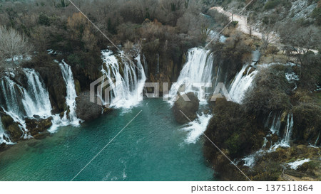 Aerial horizontal view of Kravica Waterfalls in Bosnia during winter, with powerful cascades flowing into a turquoise pool, surrounded by bare trees and rocky cliffs in a serene natural landscape 137511864
