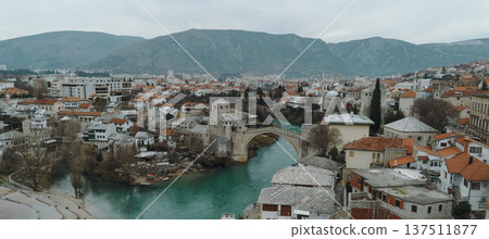Aerial view of the historic stone bridge over the Neretva River in Mostar, with old town architecture, turquoise water, and surrounding mountains under an overcast sky. Amazing Bosnia and Herzegovina 137511877