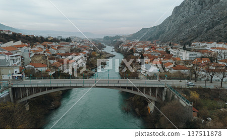 Aerial view of a road bridge crossing the Neretva River in Mostar, surrounded by residential houses with red tiled roofs, urban neighborhoods and hills under a cloudy sky Aerial view of a road bridge crossing the Neretva River in Mostar, surrounded by residential houses with red tiled roofs, urban neighborhoods and hills under a cloudy sky 137511878
