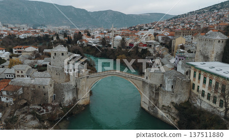 Aerial view of the historic stone bridge over the Neretva River in Mostar, with old town architecture, turquoise water, and surrounding mountains under an overcast sky. Amazing Bosnia and Herzegovina Aerial view of the historic stone bridge over the Neretva River in Mostar, with old town architecture, turquoise water, and surrounding mountains under an overcast sky. Amazing Bosnia and Herzegovina 137511880