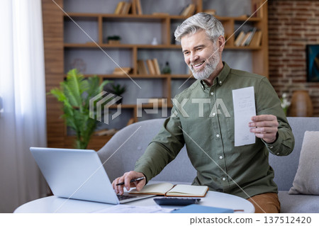 Happy senior man managing home finances, holding a paper receipt while using a laptop and notebook, demonstrating financial planning and online banking 137512420
