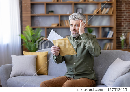 Mature man sitting on sofa at home, shocked and stressed as he opens an envelope with bad financial news unexpected bill or debt notice, hand to head, worried and upset 137512425