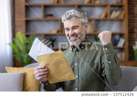 Happy senior man sitting on a sofa at home, opening an envelope and reading a letter with good news, celebrating a success or achievement with a raised fist 137512426