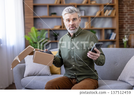 Mature man sitting on sofa opening a small cardboard box containing a damaged product, holding a smartphone, looking shocked and frustrated with the delivery issue 137512459
