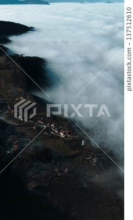Aerial view of a small mountain village partially covered by low clouds, with houses, fields and forested slopes emerging from fog in soft morning light. Amazing Bosnia and Herzegovina 137512610