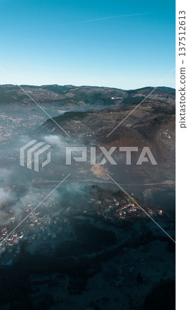 Aerial view of a small mountain village partially covered by low clouds, with houses, fields and forested slopes emerging from fog in soft morning light. Amazing Bosnia and Herzegovina Aerial view of a small mountain village partially covered by low clouds, with houses, fields and forested slopes emerging from fog in soft morning light. Amazing Bosnia and Herzegovina 137512613