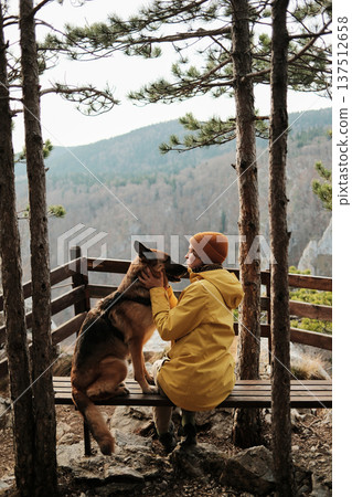 A woman in a yellow jacket sits on a bench in Tara National Park holding a German Shepherd. Concept of emotional bond travel and hiking with pets in wild mountain nature 137512658