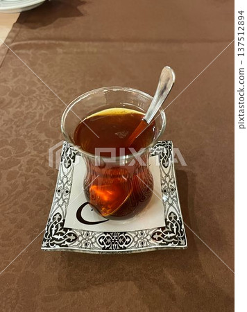 Traditional Turkish tea served in a small glass with a spoon on a decorative saucer. Warm beverage scene emphasizing culture, hospitality, and calm everyday ritual Traditional Turkish tea served in a small glass with a spoon on a decorative saucer. Warm beverage scene emphasizing culture, hospitality, and calm everyday ritual 137512894