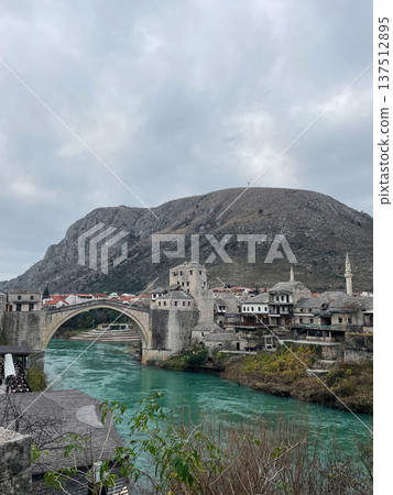 View of the historic Stari Most bridge in Mostar with stone houses, turquoise river, and mountain backdrop. Iconic Balkan landmark showing medieval architecture and heritage View of the historic Stari Most bridge in Mostar with stone houses, turquoise river, and mountain backdrop. Iconic Balkan landmark showing medieval architecture and heritage 137512895