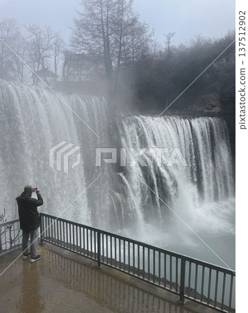 Tourist photographing Pliva Waterfall in Jajce, Bosnia and Herzegovina, standing on viewpoint platform with powerful cascades, mist and dramatic winter atmosphere Tourist photographing Pliva Waterfall in Jajce, Bosnia and Herzegovina, standing on viewpoint platform with powerful cascades, mist and dramatic winter atmosphere 137512902