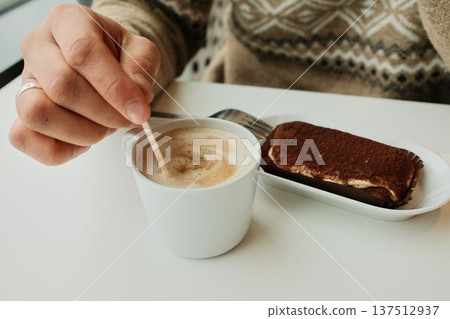 A hand stirs foamy cappuccino in a white cup beside a tiramisu dessert served on a plate in a minimalist cafe setting 137512937