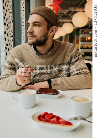 A man sits at a cafe table with cappuccino and desserts, holding a fork and looking away from the window in a cozy modern interior A man sits at a cafe table with cappuccino and desserts, holding a fork and looking away from the window in a cozy modern interior 137512938