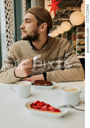 A man sits at a cafe table with cappuccino and desserts, holding a fork and looking away from the window in a cozy modern interior 137512940