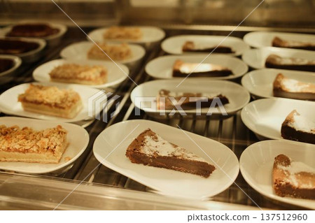 Assorted cake slices arranged on white plates inside a bakery display case. Sweet dessert selection showcasing variety, freshness and cafe pastry menu 137512960