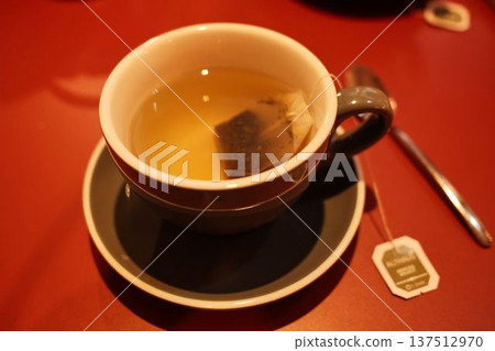 Hot tea in a ceramic cup with teabag resting inside, placed on a red table. Warm indoor light creates a cozy atmosphere of relaxation and quiet evening comfort 137512970