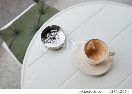 Empty coffee cup on a white table with an ashtray nearby. Minimal urban scene reflecting cafe routine, pause, habits, and everyday lifestyle atmosphere 137512974