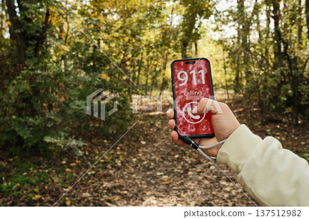 Hand holding a smartphone showing a 911 emergency call screen in a forest path. Concept of danger, urgent help request, outdoor accident risk and emergency communication in nature 137512982